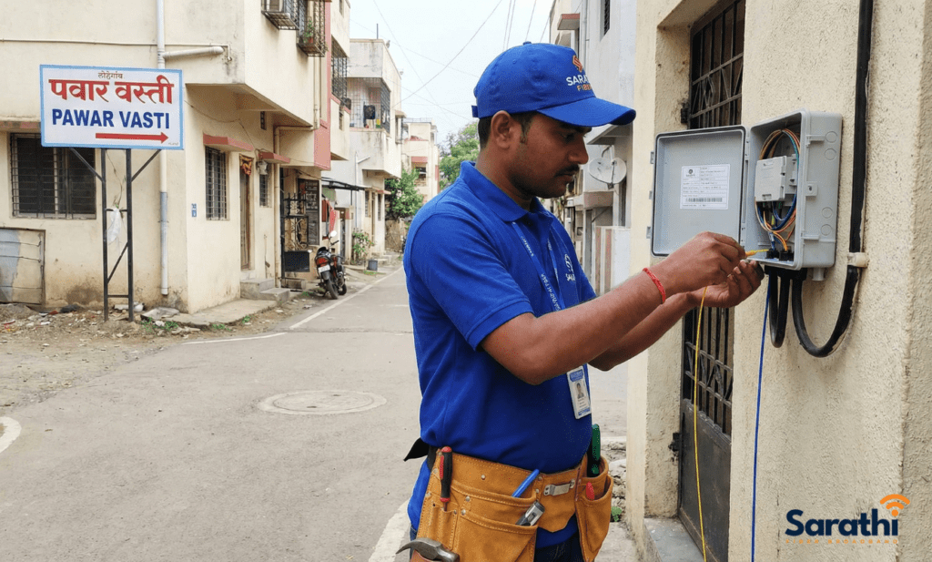 A Sarathi Fiber technician in a blue uniform and cap working on a fiber optic junction box in Pawar Vasti, Lohegaon.