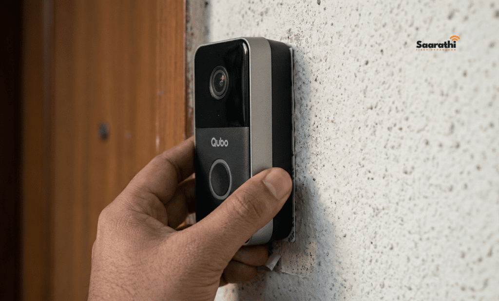 A macro close-up photograph focused on the mounting side of a battery-powered Qubo Smart Doorbell, showing adhesive tape being pressed against a rough painted exterior wall.