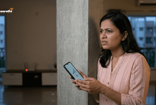 A frustrated Indian woman near a concrete structural pillar in a modern Wagholi apartment looks at her smartphone showing zero signal bars. A basic blinking red generic router is far away, visually triggering a search for how to boost WiFi signal.