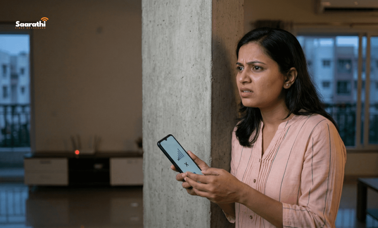 A frustrated Indian woman near a concrete structural pillar in a modern Wagholi apartment looks at her smartphone showing zero signal bars. A basic blinking red generic router is far away, visually triggering a search for how to boost WiFi signal.